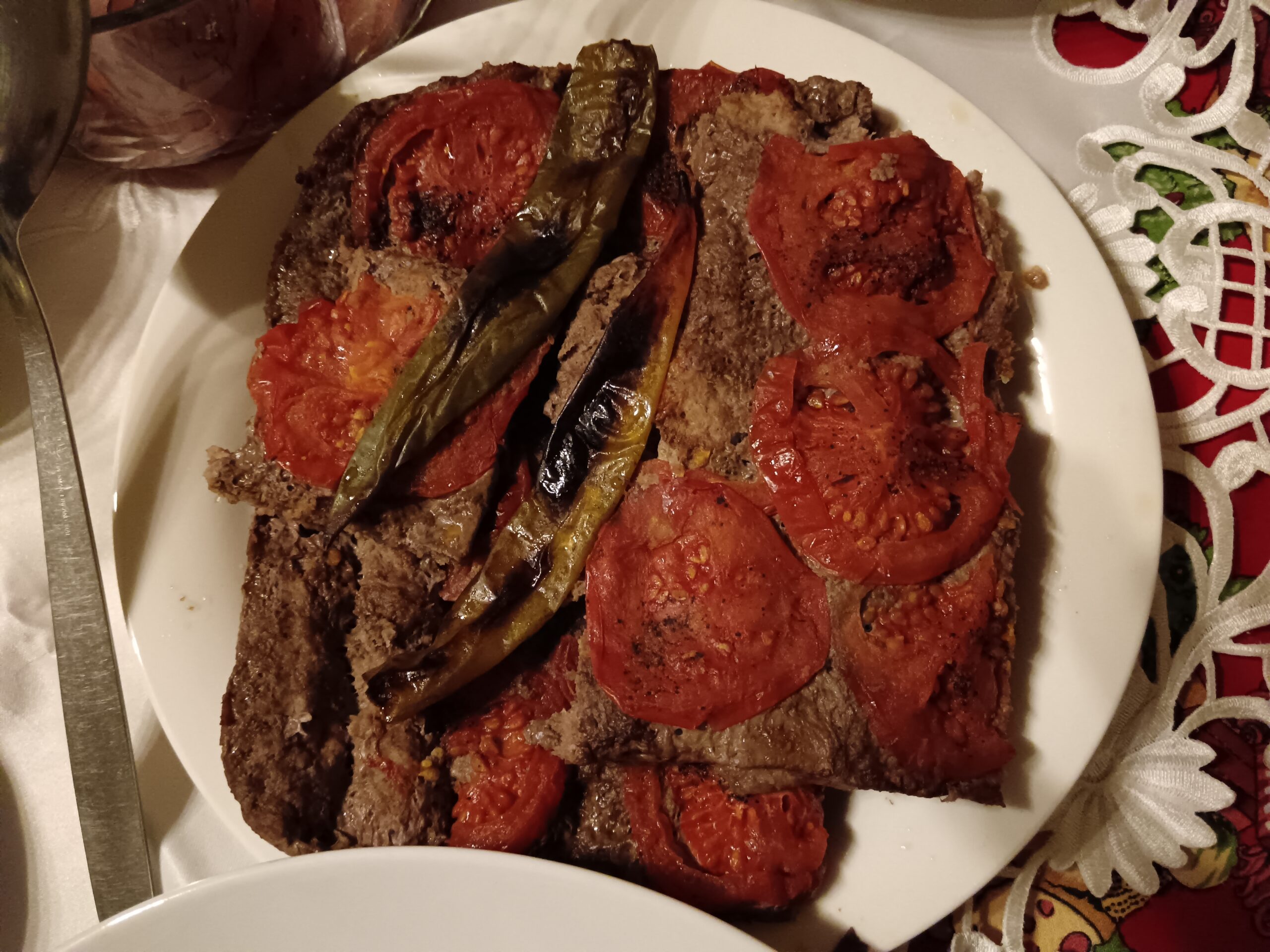 Мясо по-сирийски с запечёнными помидорами и жареным перцем: подано на белой тарелке, рядом — свежая зелень и след пальца — бесплатно скачайте фото.